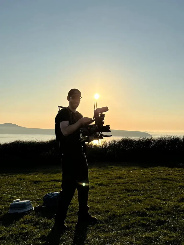 Camera Operator holding a professional camera. He's looking into camera and the sun is behind him. He's quite silhouetted but not entirely. It's a beautiful day and the sun is low in the sky. It's a still shot from a brand video production for a US based silicon valley tech startup called Hipcamp.