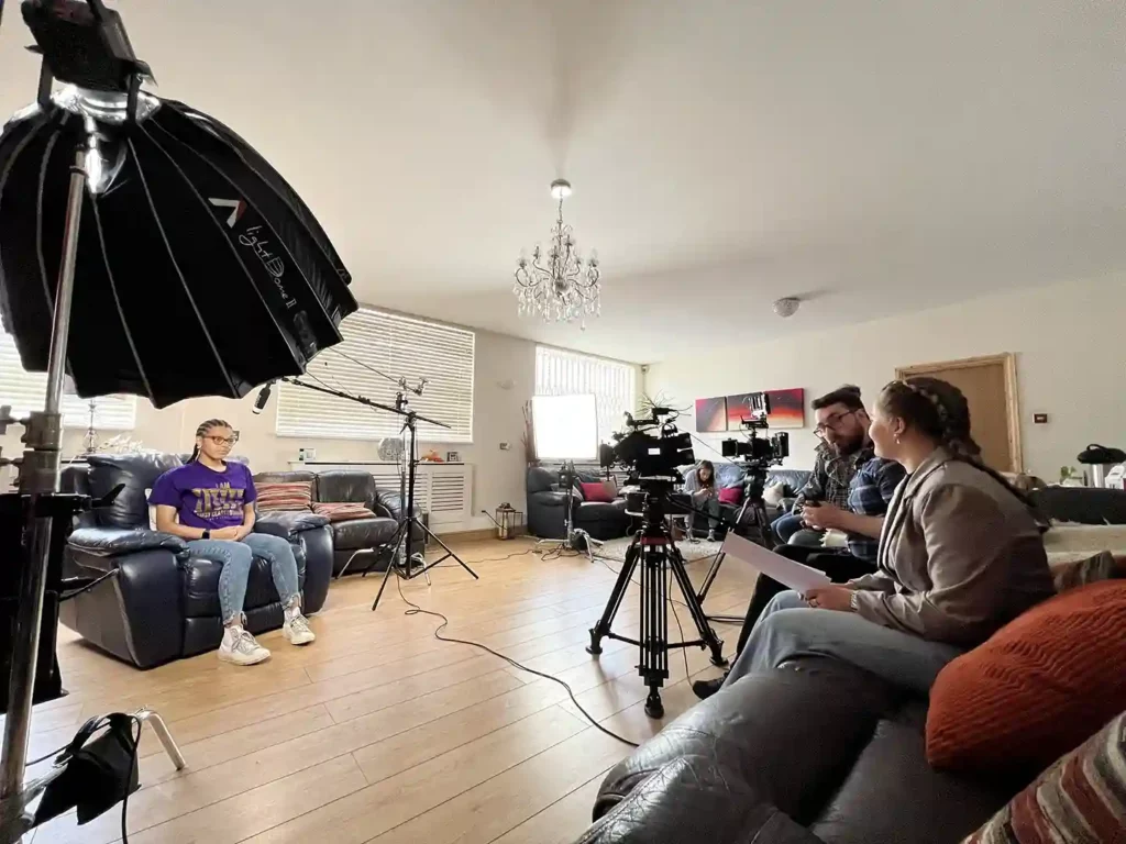 Charity Video Case Study featured image features a young woman being interviewed by a young female producer. A camera operator sits nearby observing. There are two professional video cameras and a lot of lighting.