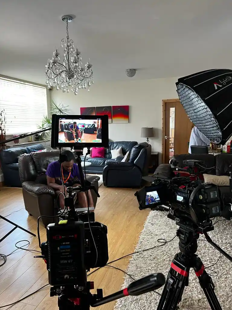 A behind the scenes image from a video shoot for a charity in Birmingham, England. There is a load of camera equipment in the foreground and a young black man sitting on an armchair waiting to be interviewed about his work.