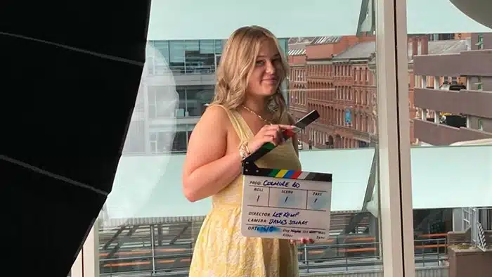 An image of the young woman who is the subject of the blog post. She is standing in a meeting room slightly overshadowed by large lights. She's wearing a yellow summer dress and holding a film shoot clapperboard.