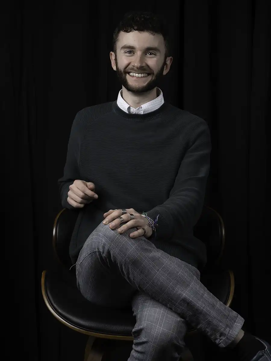 A portrait style photo of a man with a short trimmed beard. He's seated on a stool and is looking slightly off camera in a wryly amused fashion.